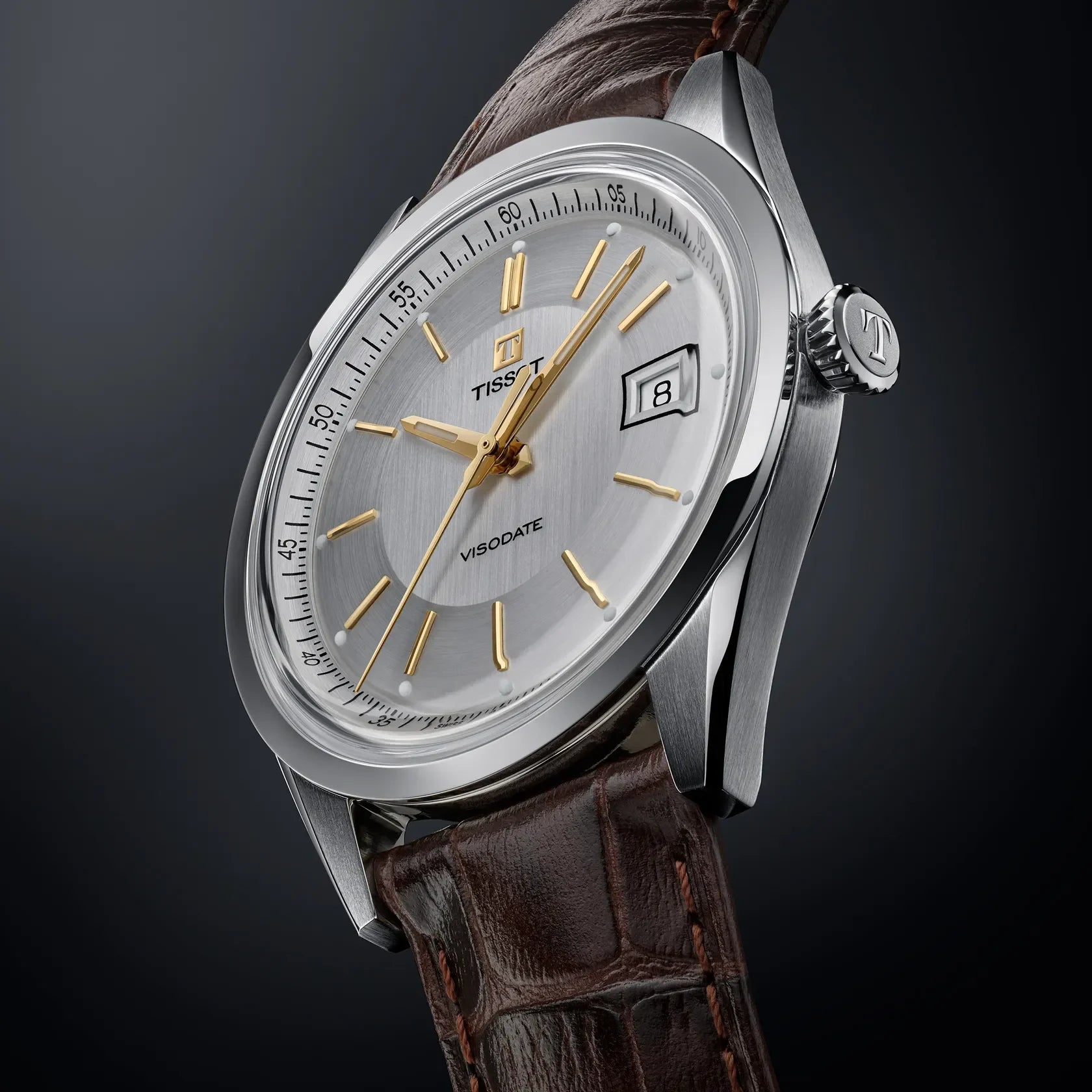 Schräge Nahaufnahme der Tissot Heritage Visodate Herrenuhr mit silbernem Zifferblatt, goldenen Indizes und braunem Lederarmband vor dunklem Hintergrund